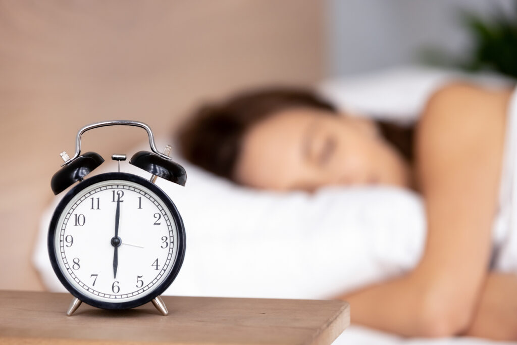 A clock placed on a table and behind it there is a girl who is sleeping